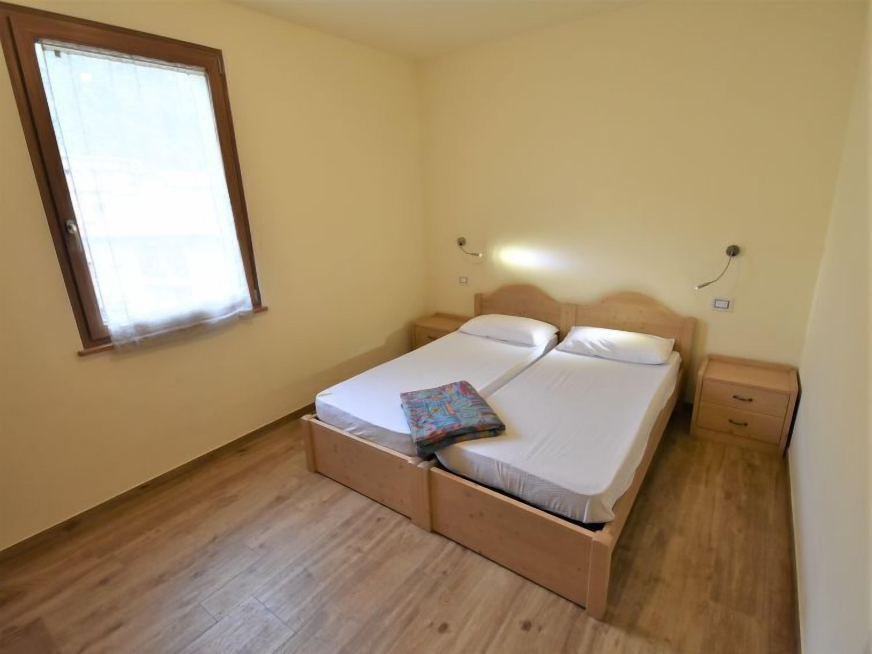 Ein Zimmer mit einem Einzelbett, Holzrahmen, einem Fenster mit Jalousien, einem Nachttisch und einem Beistelltisch.
