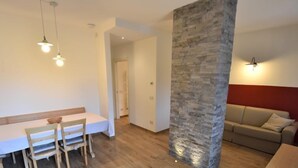 Un salon moderne avec un plancher en bois, un pilier en pierre, une table à manger blanche avec des chaises et un canapé beige.