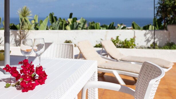 Un tavolo bianco con due bicchieri e fiori rossi, sedie in vimini bianco e vista sul mare.