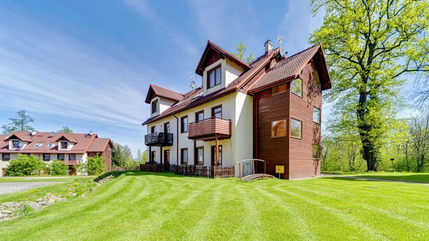 Une maison à deux étages avec une pelouse bien entretenue, une terrasse en bois et un grand arbre en arrière-plan.