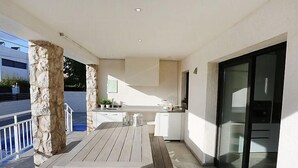 Un área de cocina al aire libre moderna con una mesa de madera y bancos, un pilar de piedra y una puerta de cristal que da a un balcón.