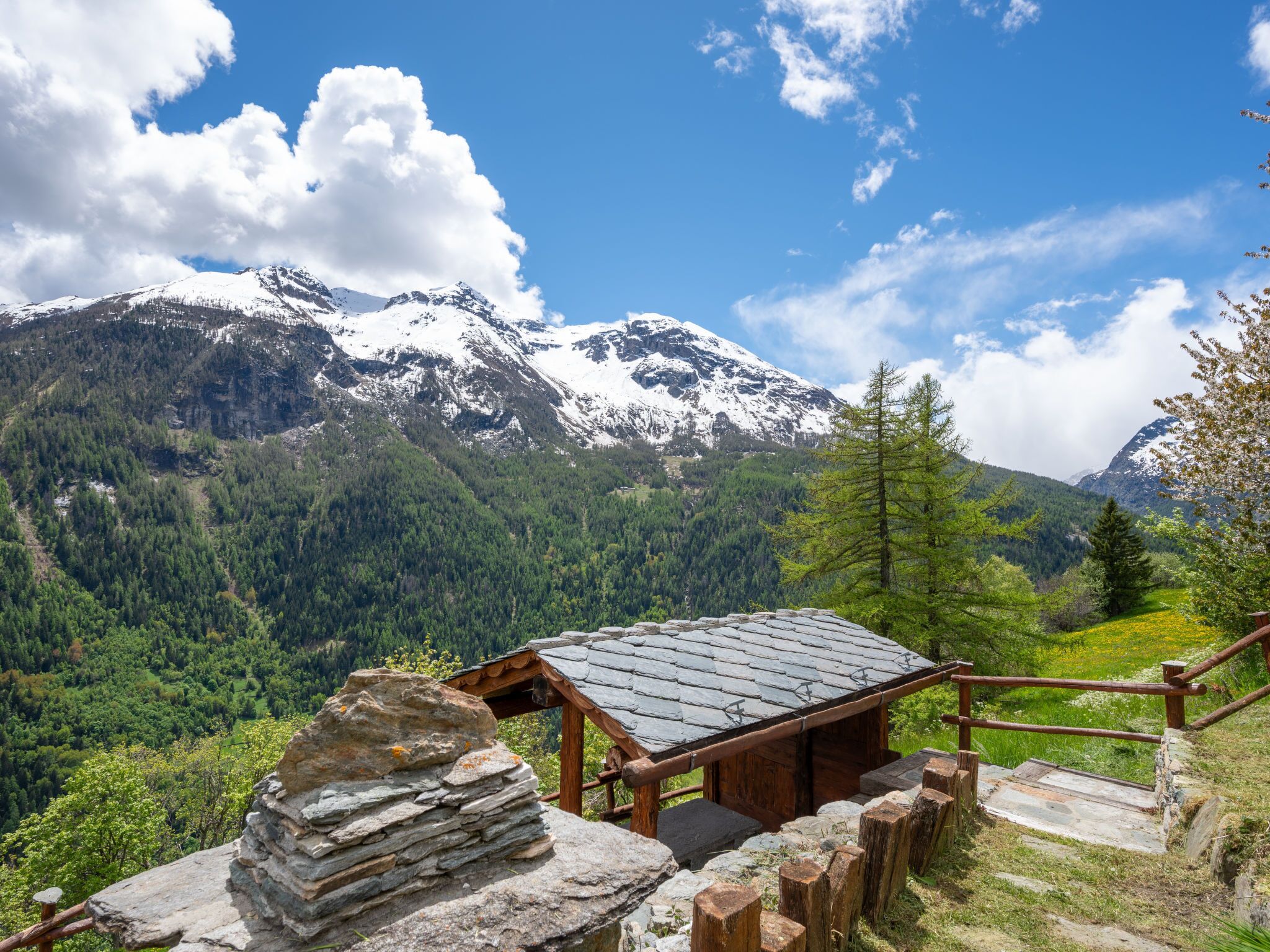 A mountain lodge with a stone staircase leading up to a wooden deck, surrounded by lush greenery and snow-capped mountains.