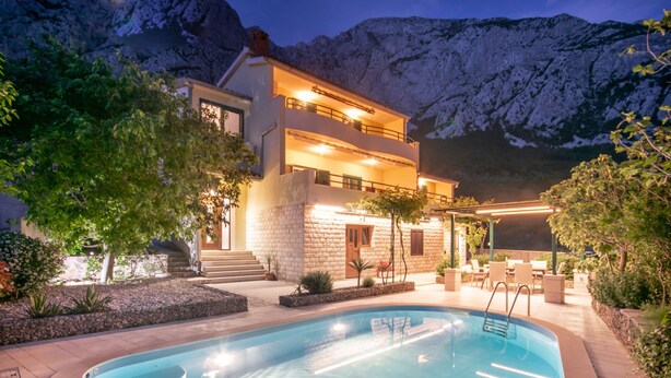 Una casa moderna de dos pisos con piscina, rodeada por un jardín ajardinado y un telón de fondo montañoso.