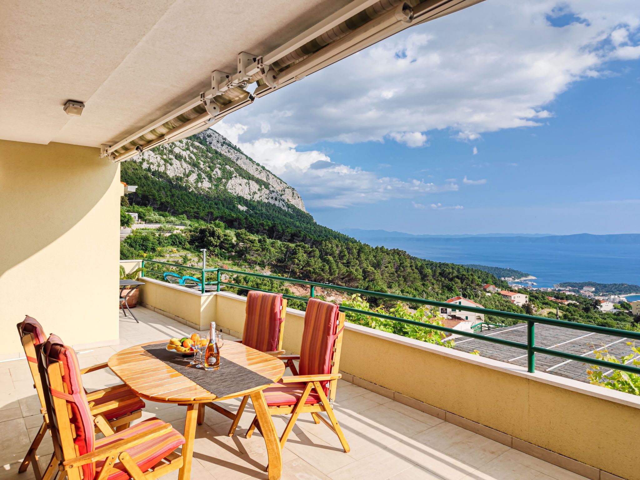 Un balcone con tavolo e sedie in legno, affacciato su un paesaggio costiero con montagne e vista mare.