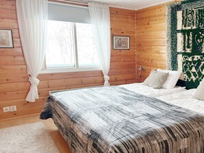 A bedroom with a large bed, wooden walls, a window with curtains, and a green patterned wall hanging.