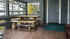 Una terraza de madera con una mesa de picnic y bancos, una puerta y grandes ventanales con vistas a un paisaje natural.