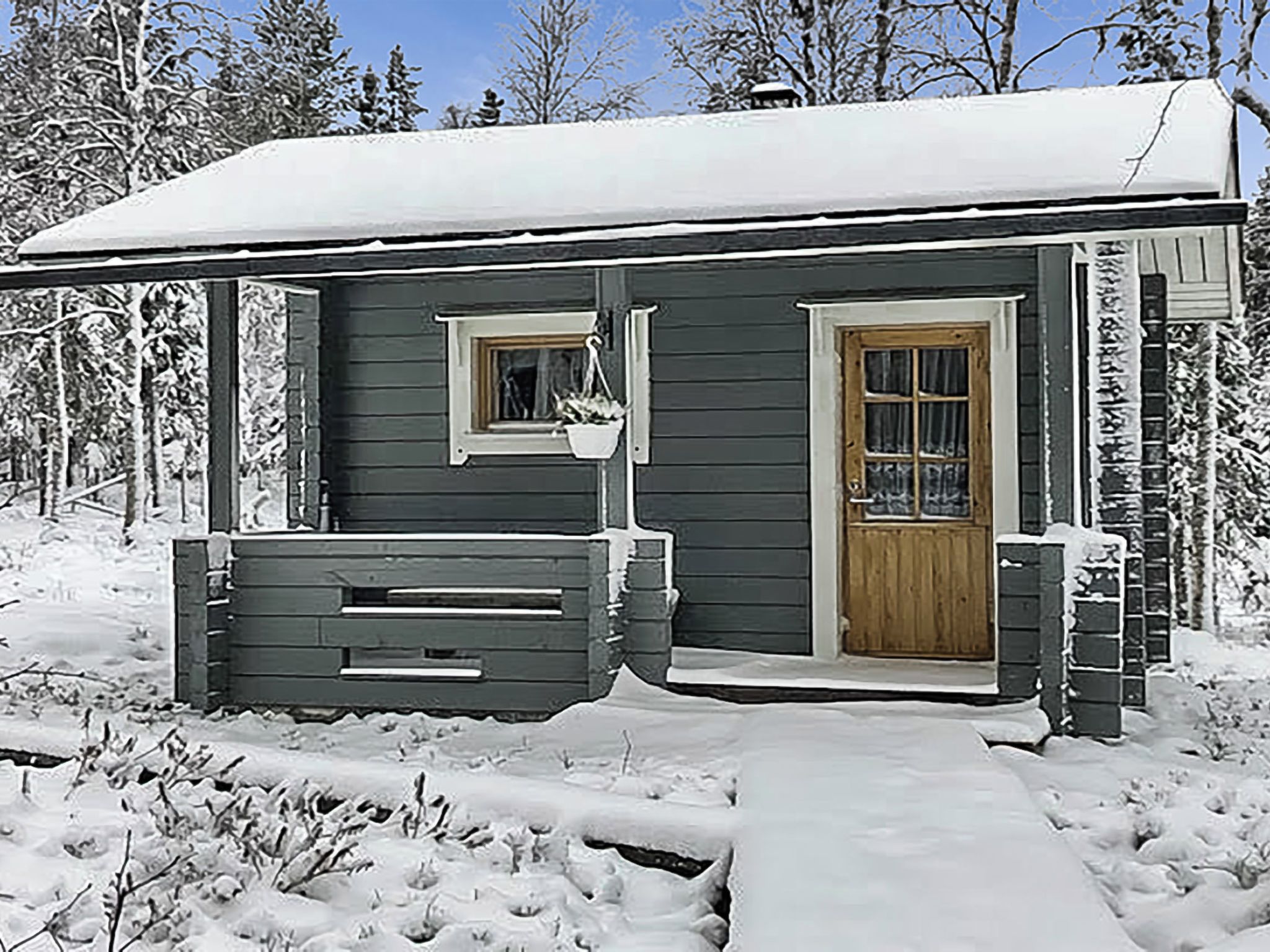 Een klein, donker houten vakantiehuisje met één deur en raam, omringd door sneeuw.