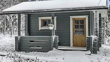 Een klein, donker houten vakantiehuisje met één deur en raam, omringd door sneeuw.