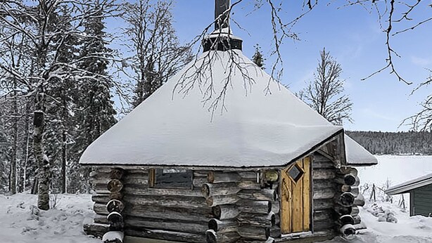 Una cabaña de troncos con techo nevado y chimenea, rodeada de nieve y árboles.