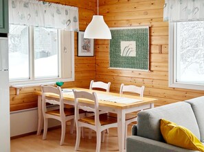 Ett mysigt rum inrett som en stuga, med träpanel på väggarna, ett matbord med stolar, en grå soffa och ett fönster med utsikt över träd.