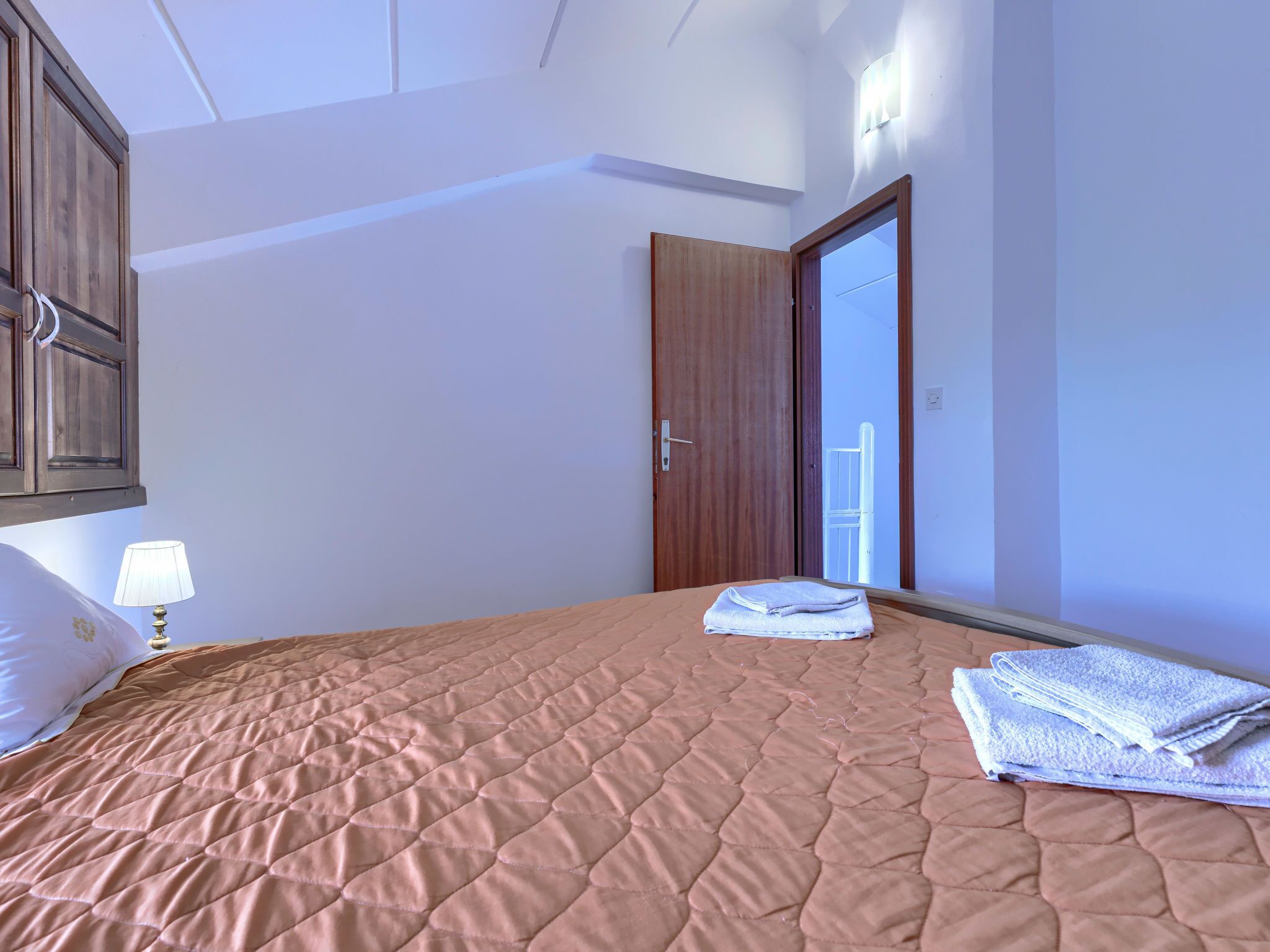 A bedroom with a bed, a nightstand, a lamp, a wooden wardrobe, and a door leading to a balcony.