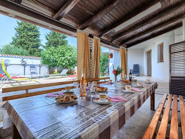 A dining area with a long table set for a meal, a view of the outdoors, and a wooden ceiling.