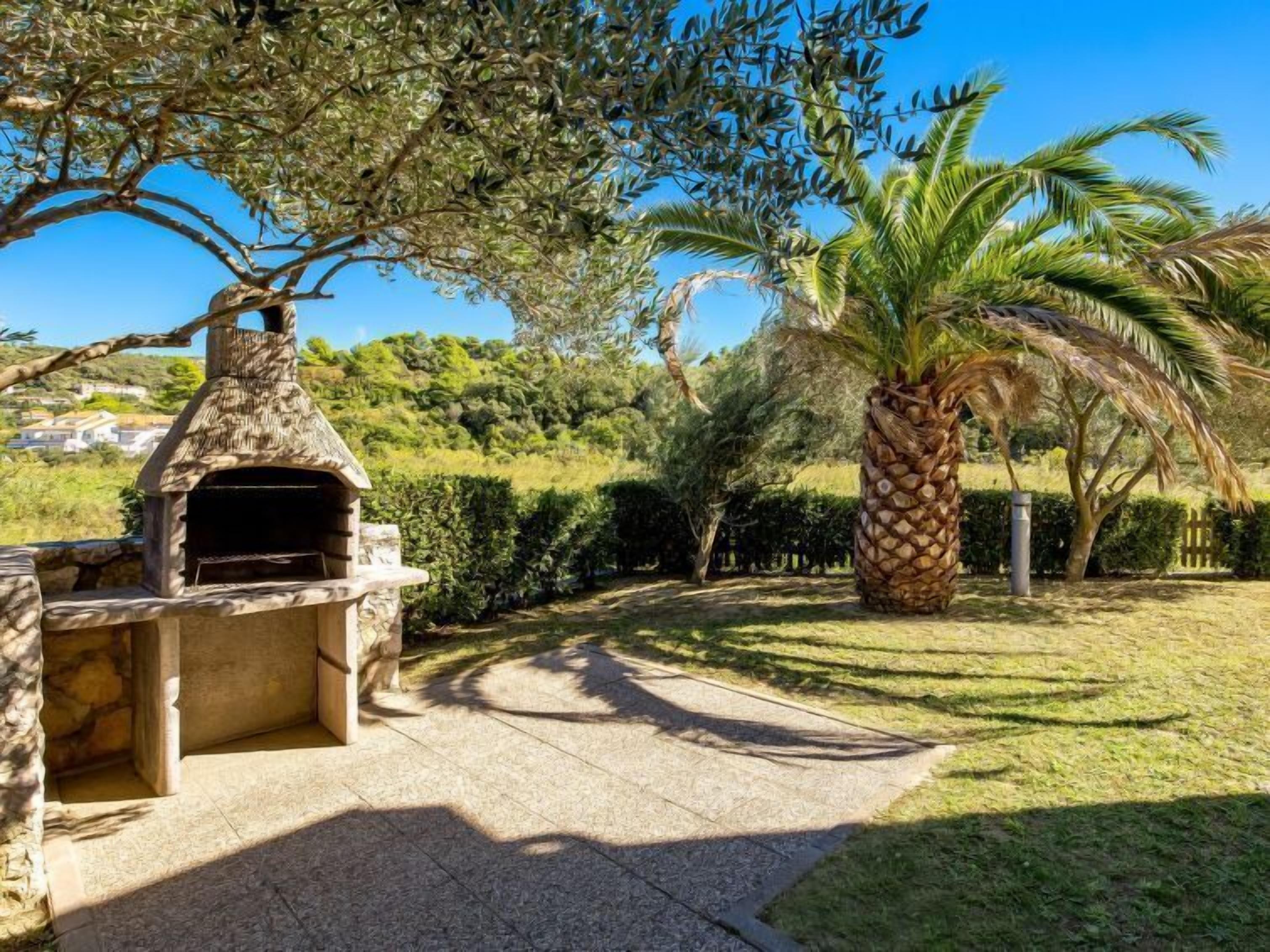 Una cocina exterior de piedra con chimenea, rodeada de setos cuidados y palmeras.