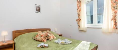 A neatly arranged bedroom with a bed, a bedside table, a lamp, a window with curtains, and a small picture on the wall.