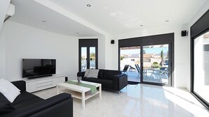 Ein modernes Wohnzimmer mit Flachbildfernseher, schwarzen Sofas, einem Couchtisch und großen Schiebeglastüren, die auf eine Terrassenfläche im Freien führen.