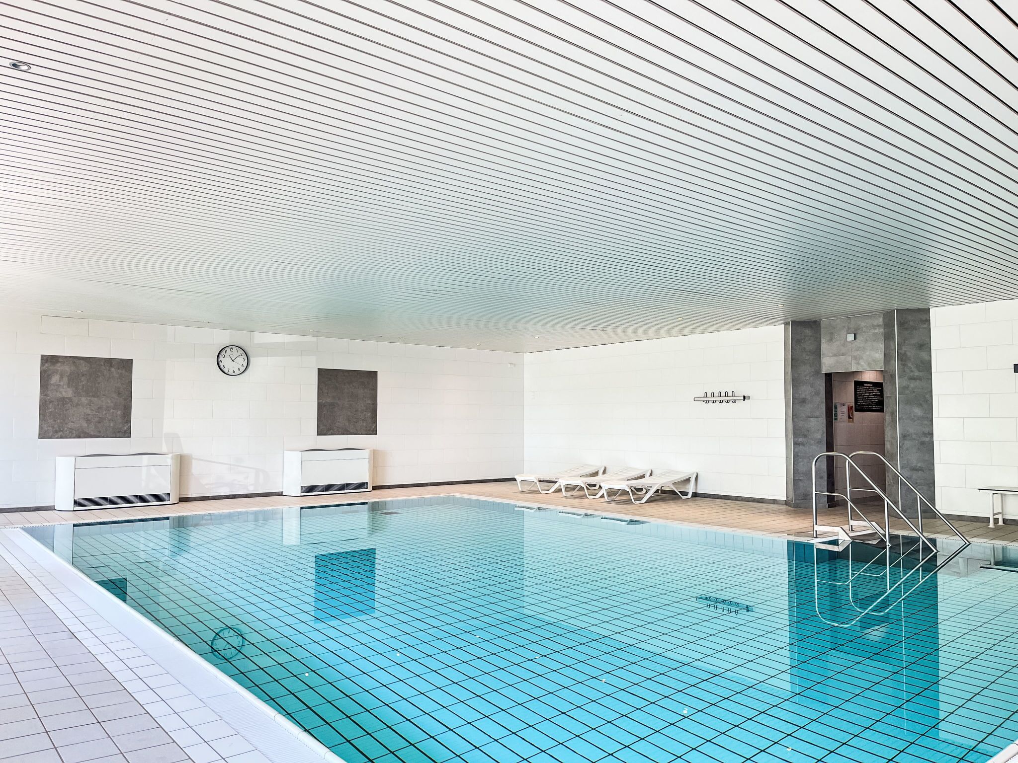 A swimming pool with tiled walls and ceiling, lounge chairs, and a clock on the wall.