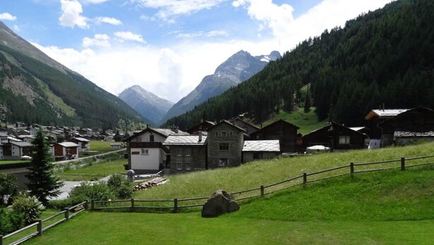 A mountain village with traditional wooden houses, a grassy foreground, and a river.