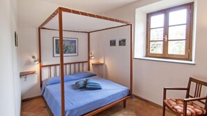 A four-poster bed with a blue bedspread and pillows, a wooden chair with a cushion, a small table with a lamp, and a window with a view of buildings. - I Tigli by Interhome (Rufina)