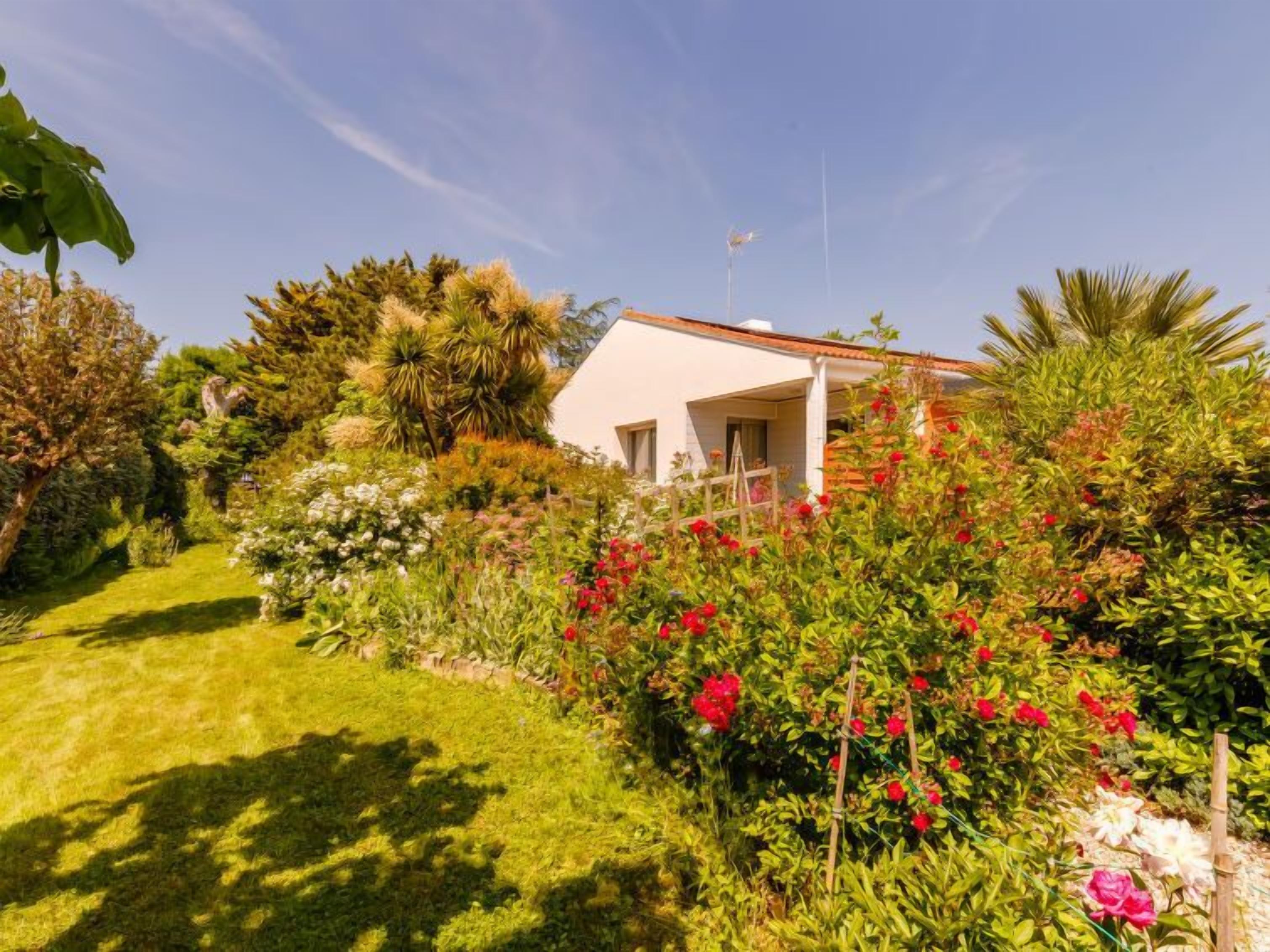 A white house with a sloped roof, surrounded by a lush garden with various plants and flowers.