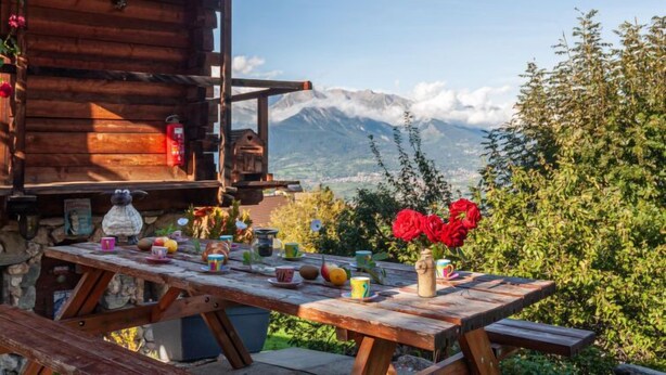 A wooden cabin with a table set for a meal, surrounded by greenery and a mountain view. - Le Dahu by Interhome (Nendaz)