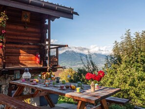 A wooden cabin with a table set for a meal, surrounded by greenery and a mountain view.