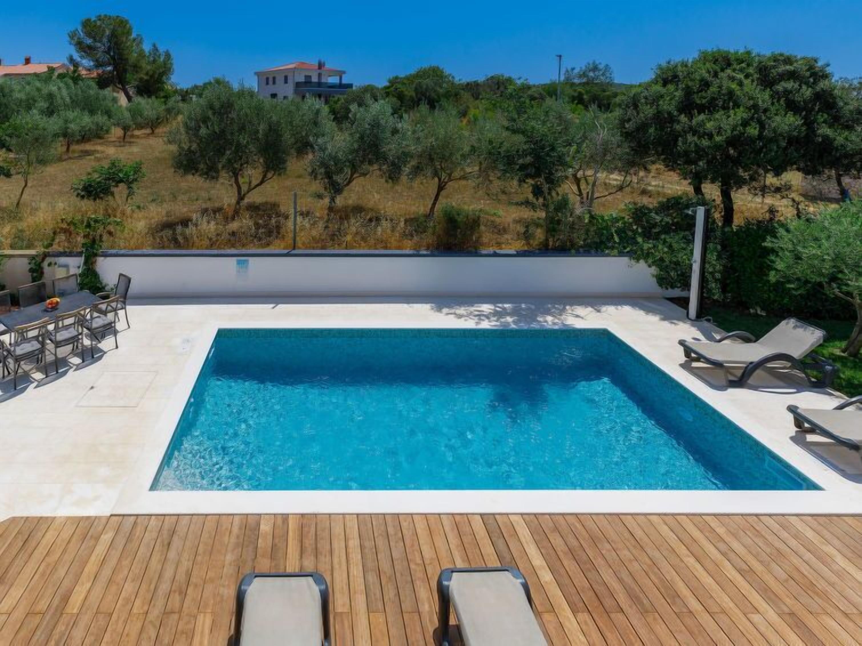Ein rechteckiger Pool mit Holzdeck und Liegestühlen, umgeben von Grünflächen und einer Hügelsicht.