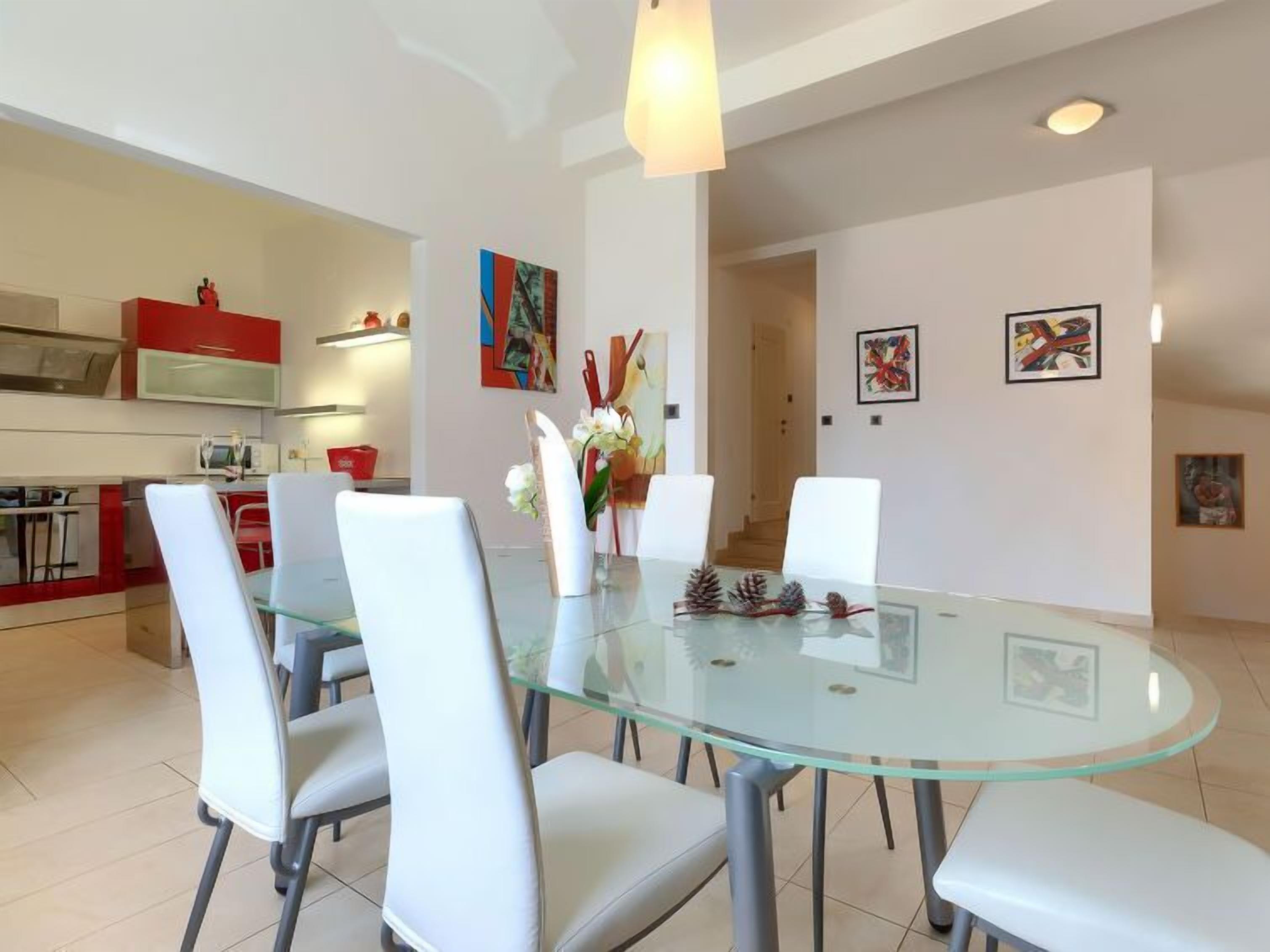 Un comedor moderno con una mesa de vidrio rodeada de sillas blancas, una cocina al fondo y obras de arte en las paredes.