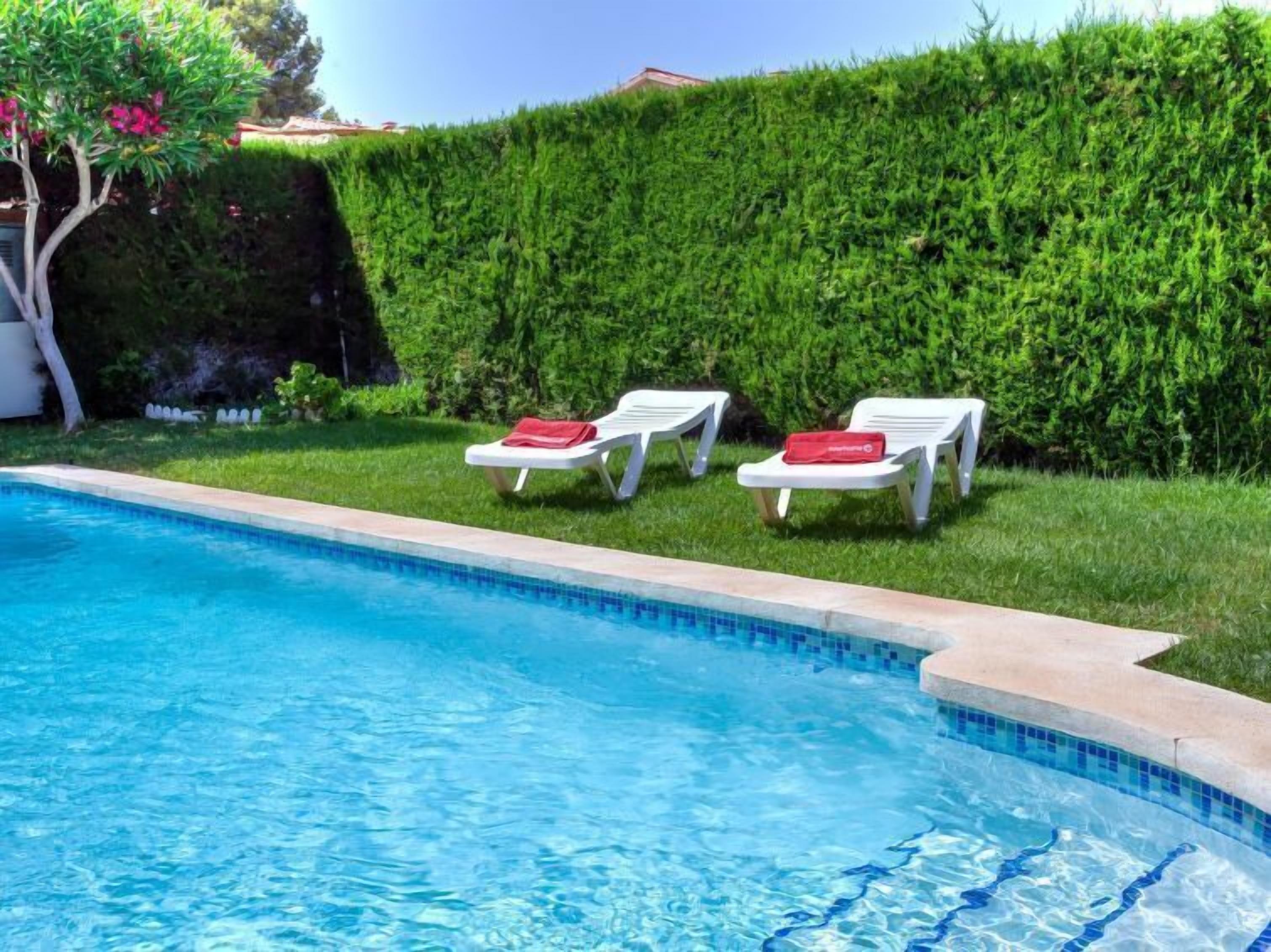 A swimming pool with two white lounge chairs and red towels, surrounded by a neatly trimmed hedge.
