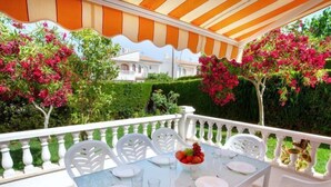 Ein Tisch für vier Personen, mit weißen Stühlen, einer Glastischplatte und einer Obstschale auf einem Balkon mit weißer Brüstung, umgeben von Grünpflanzen und rosa Blüten.