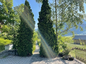 Un chemin pavé, bordé de grands arbres à feuilles persistantes, mène à un bâtiment doté d’un toit en pente.
