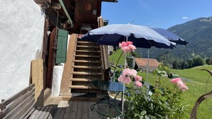 Una escalera de madera que lleva a una terraza de madera con un paraguas azul, una silla verde y flores rosadas.