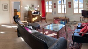 Una sala de estar acogedora con un sofá de cuero oscuro, una mesa de centro de vidrio y un tapete rojo estampado. Hay un mueble de madera con cajones y una mesita de noche con una lámpara. La habitación tiene ventanas amplias con cortinas rojas y vistas al exterior.