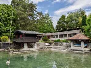 A building with a wooden deck and a small pavilion by a clear water body, surrounded by dense trees.