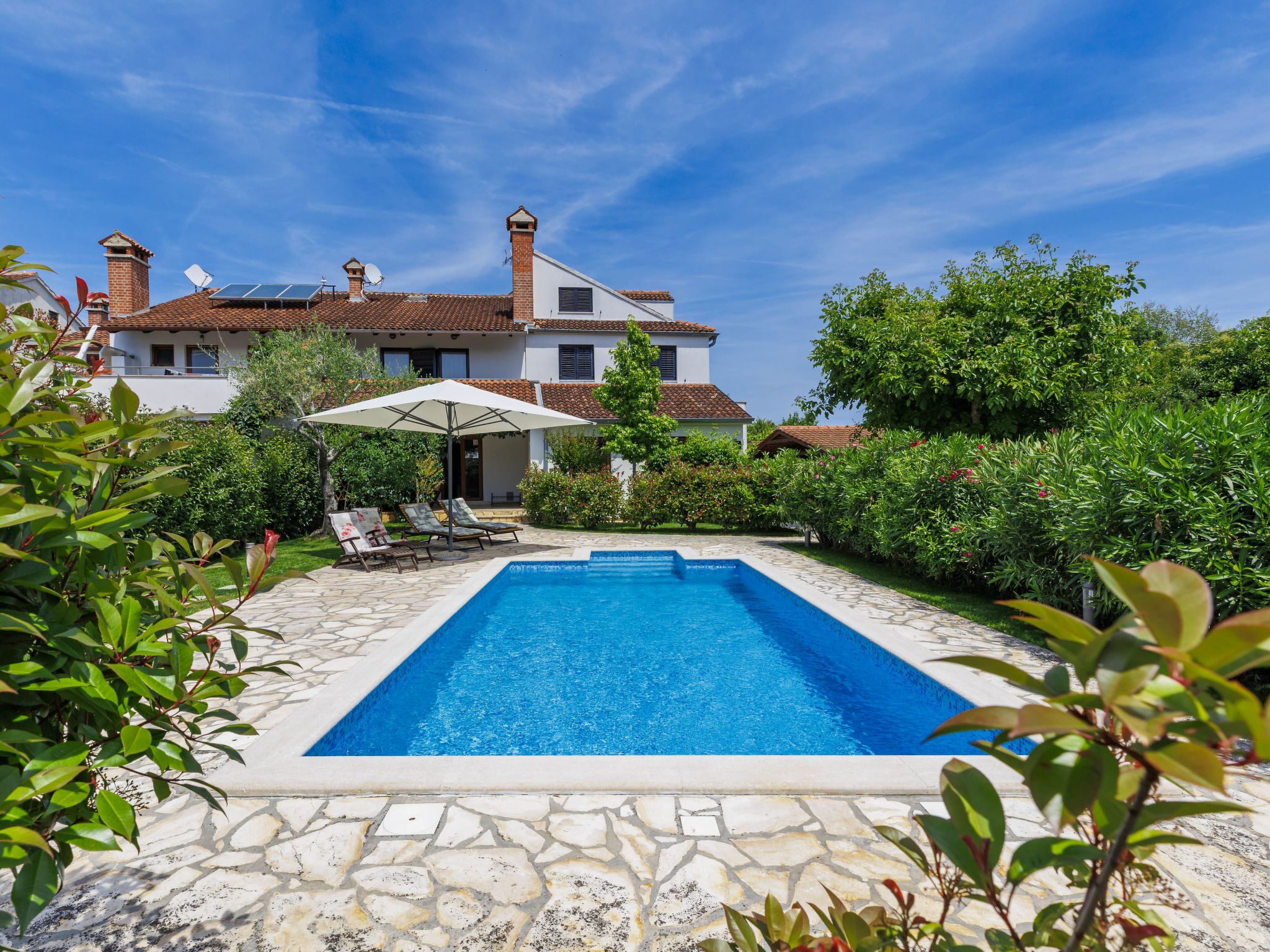 A swimming pool with stone tiles, surrounded by lush greenery and a white building with a tiled roof.