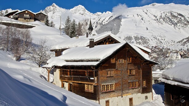 Eine Holzchalet mit einem Balkon, umgeben von schneebedeckten Bergen und einem klaren Himmel.
