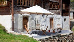 Ein rustikales Gebäude mit weißer Fassade, Holztüren und -fenstern. Ein weißer Sonnenschirm spendet Schatten, und mehrere Holztische und -stühle stehen draußen bereit.