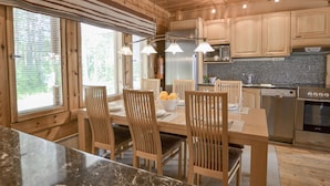 A wooden cabin interior with a dining area, kitchen, and large windows. - Kannelpirtti 2 by Interhome (Liperi)