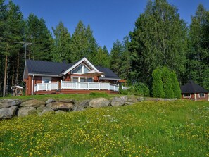 Una cabaña con terraza, rodeada por un paisaje natural con árboles y un campo de flores amarillas.