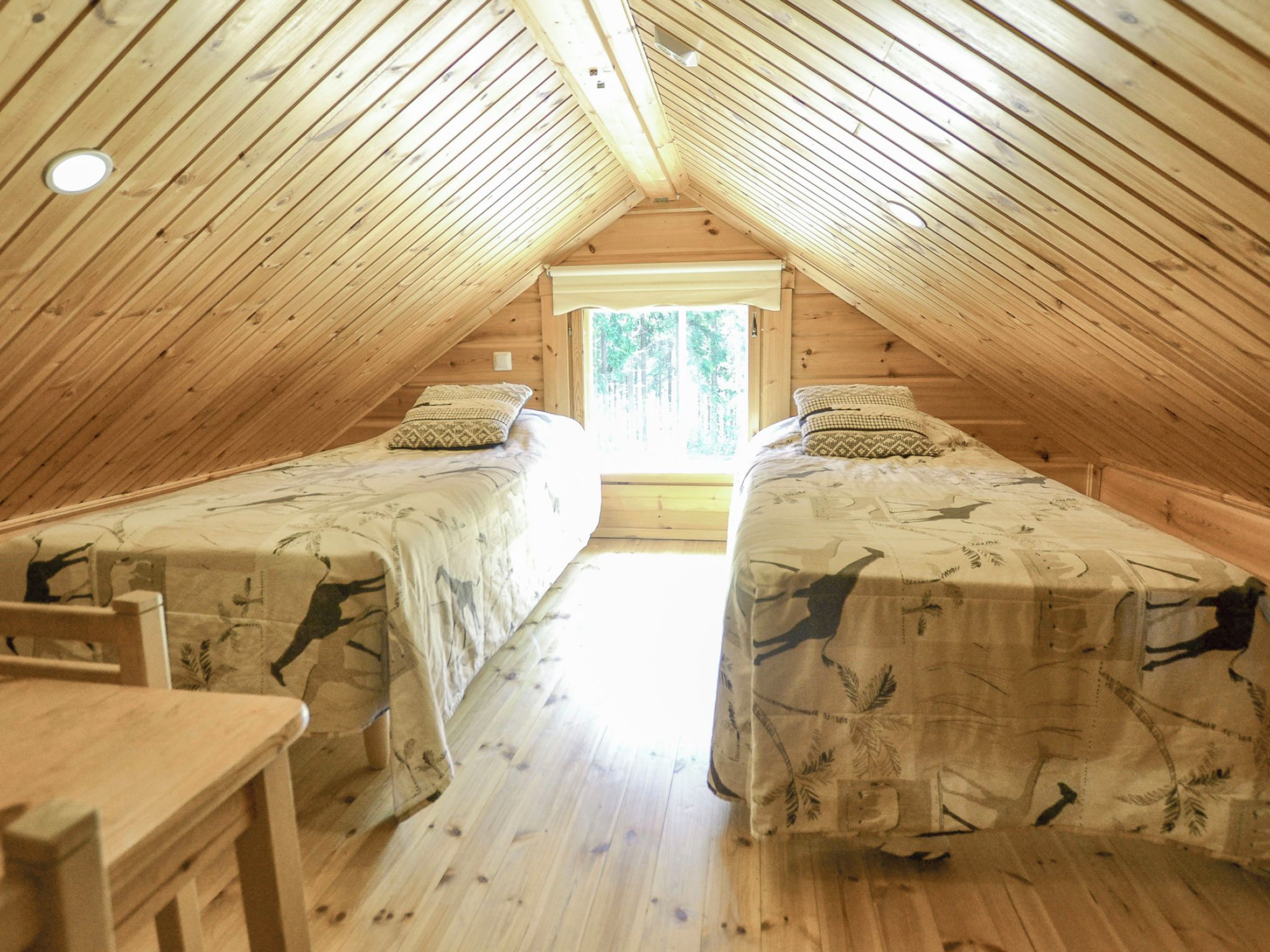 A cozy attic bedroom with two beds, a wooden desk, and a window providing a view of trees.