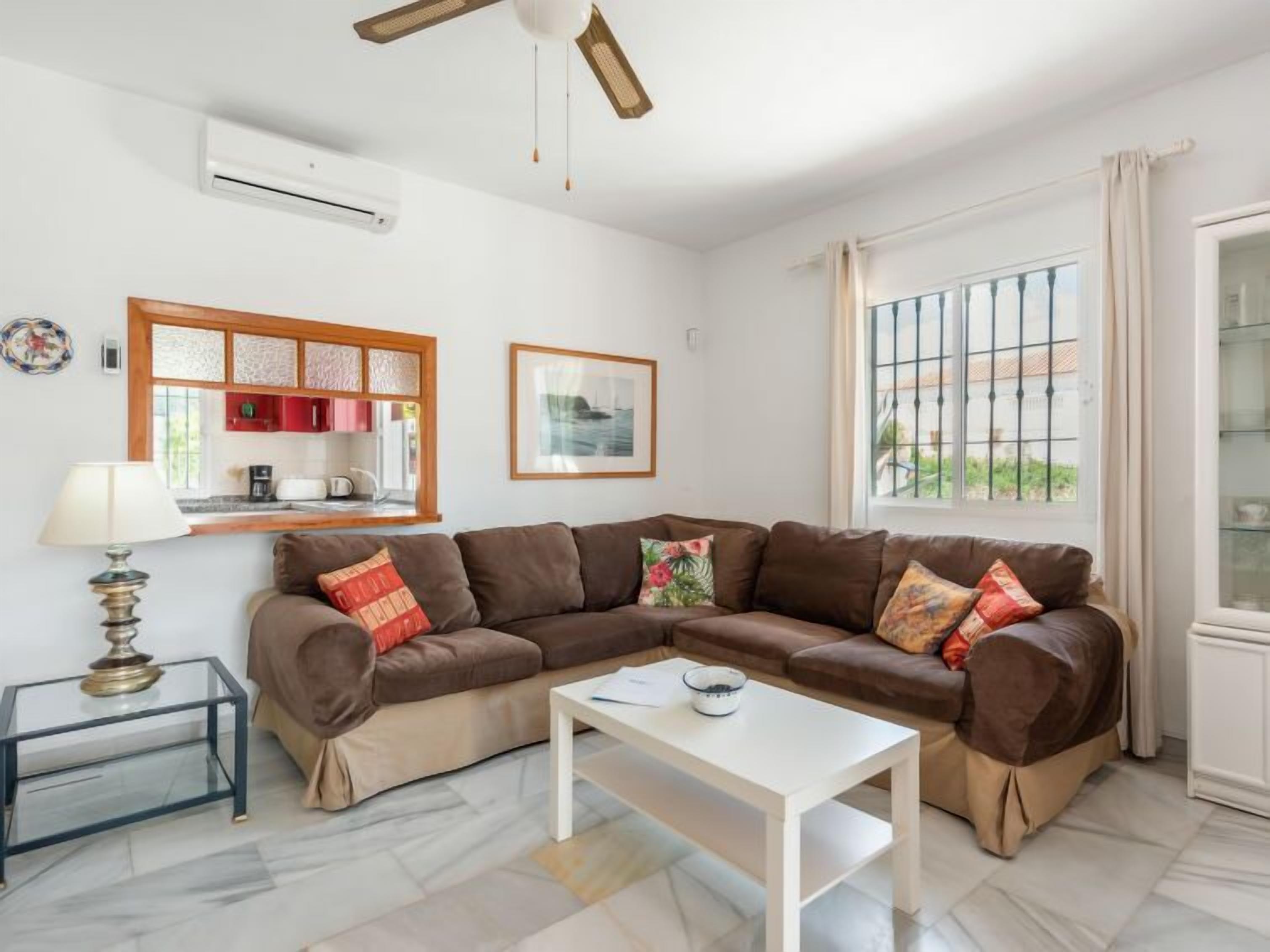 A living room with a brown sectional sofa, a white coffee table, a glass side table, a lamp, a kitchen area with red cabinets, and a window with a view of greenery.