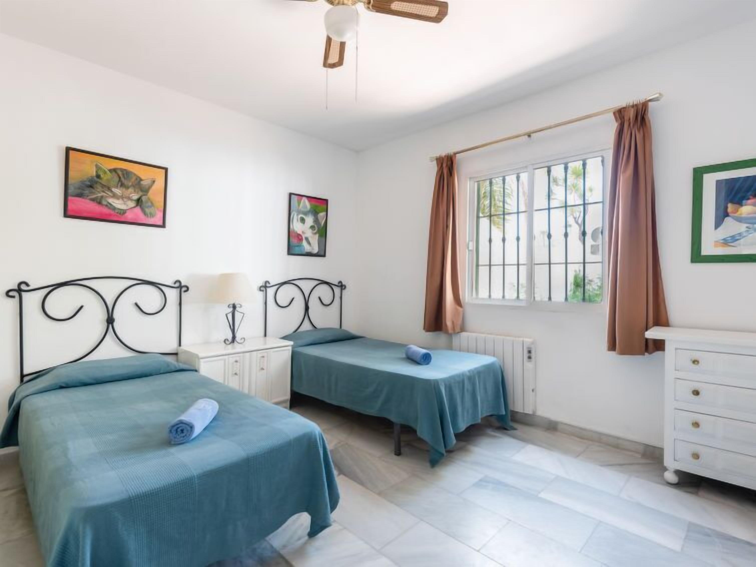 Ein Zimmer mit zwei Einzelbetten, einem weißen Schminktisch, einem Fenster mit Vorhängen und Bildern an den Wänden.
