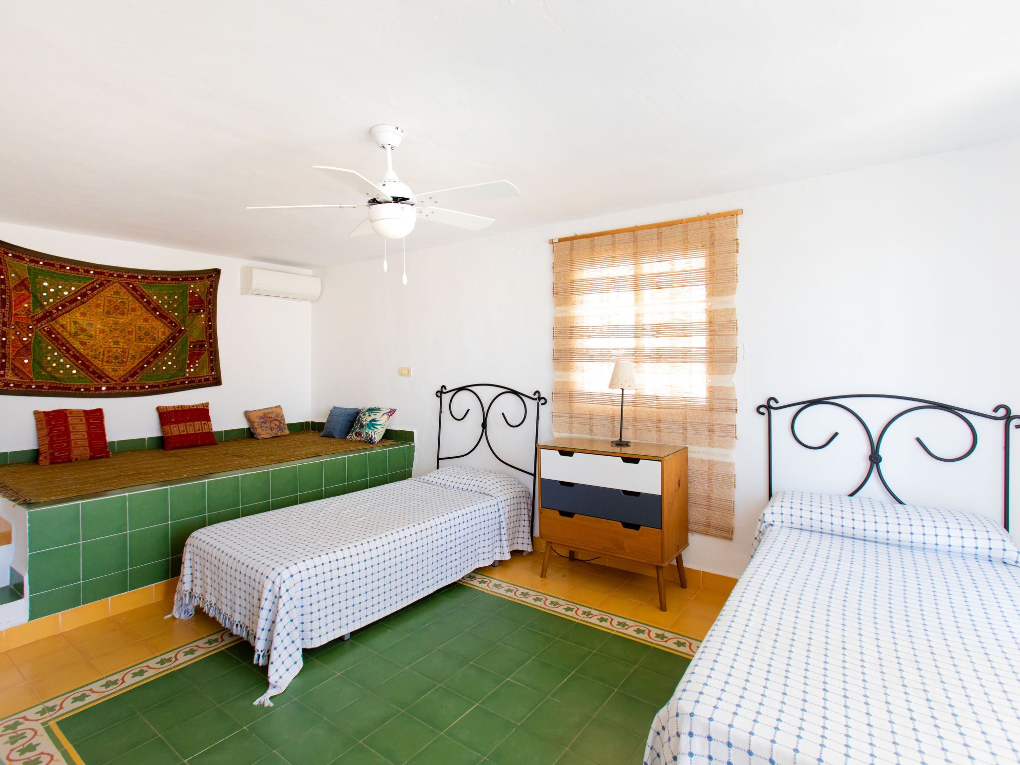 Una habitación con dos camas, un ventilador de techo, un tapiz y una ventana con persianas.