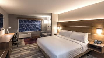 A hotel room with a large bed, a desk, a chair, a desk lamp, a wall-mounted lamp, an air conditioning unit, and a large window with a geometric pattern.