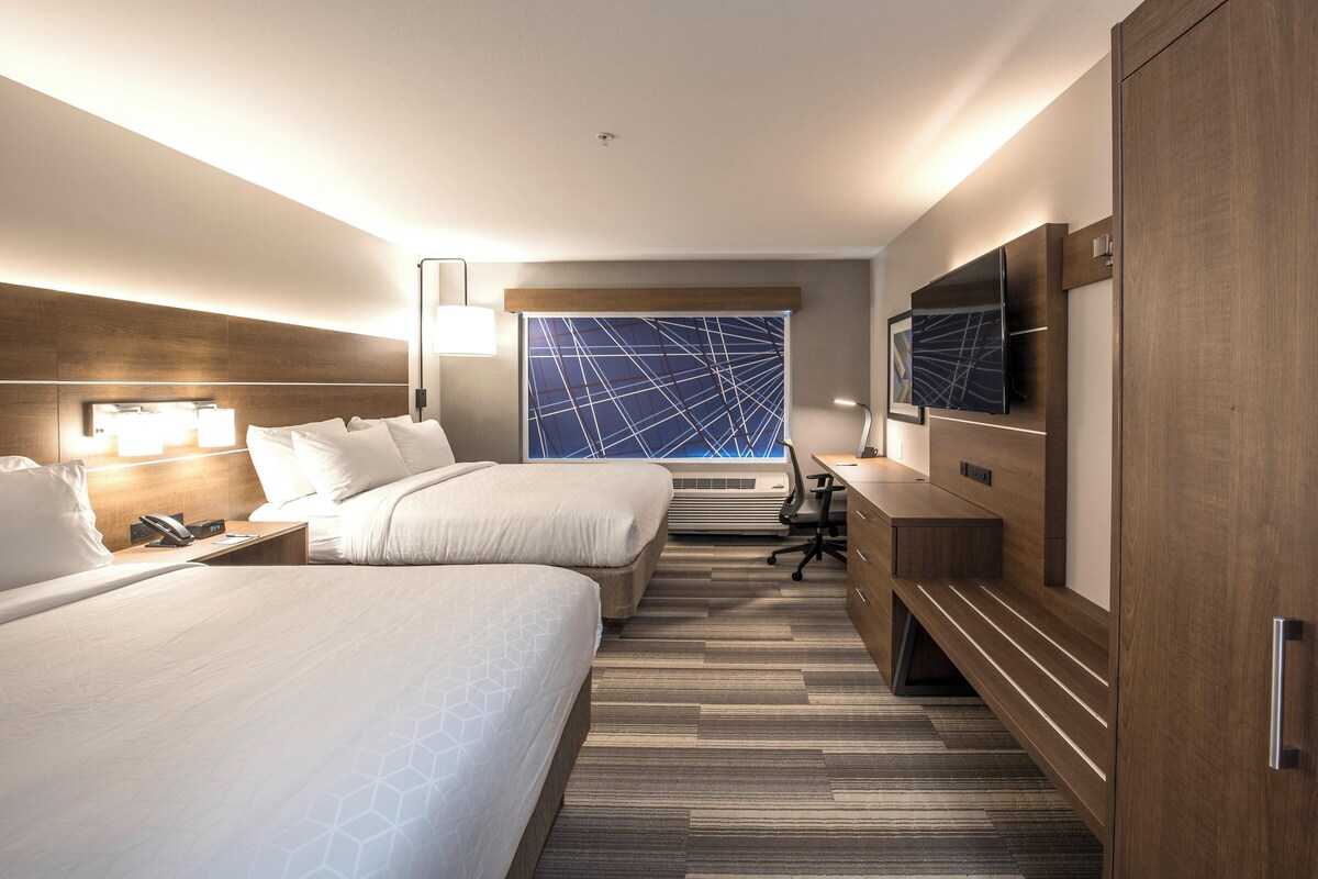 A hotel room with two beds, a desk, a TV, and a large window with a geometric view.