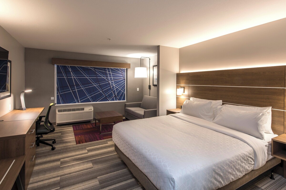 A hotel room with a large bed, a desk with a chair, a window with a geometric view, and a wall-mounted lamp.
