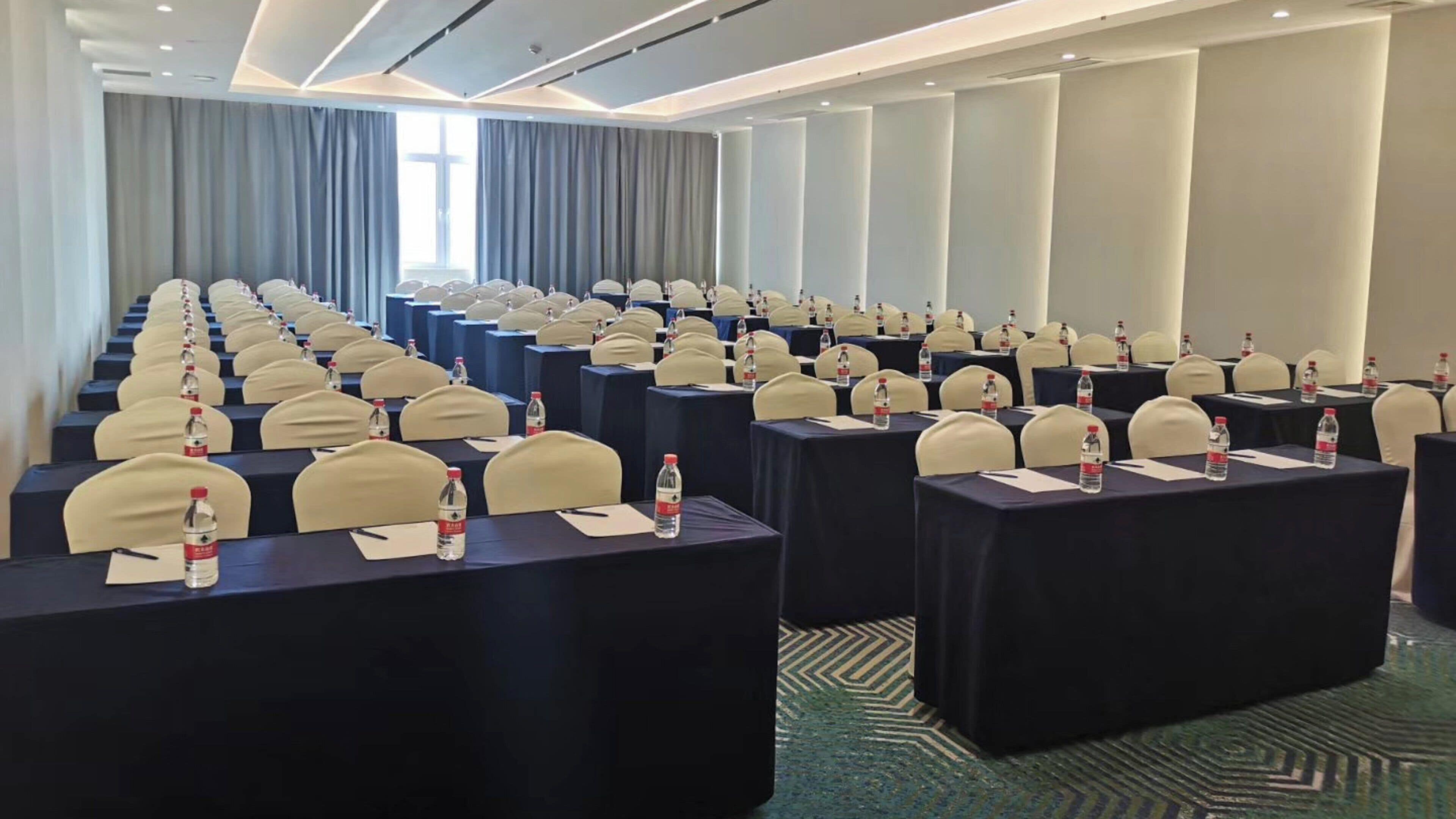 A conference room with rows of tables and chairs, water bottles on each table, and a large window with curtains.