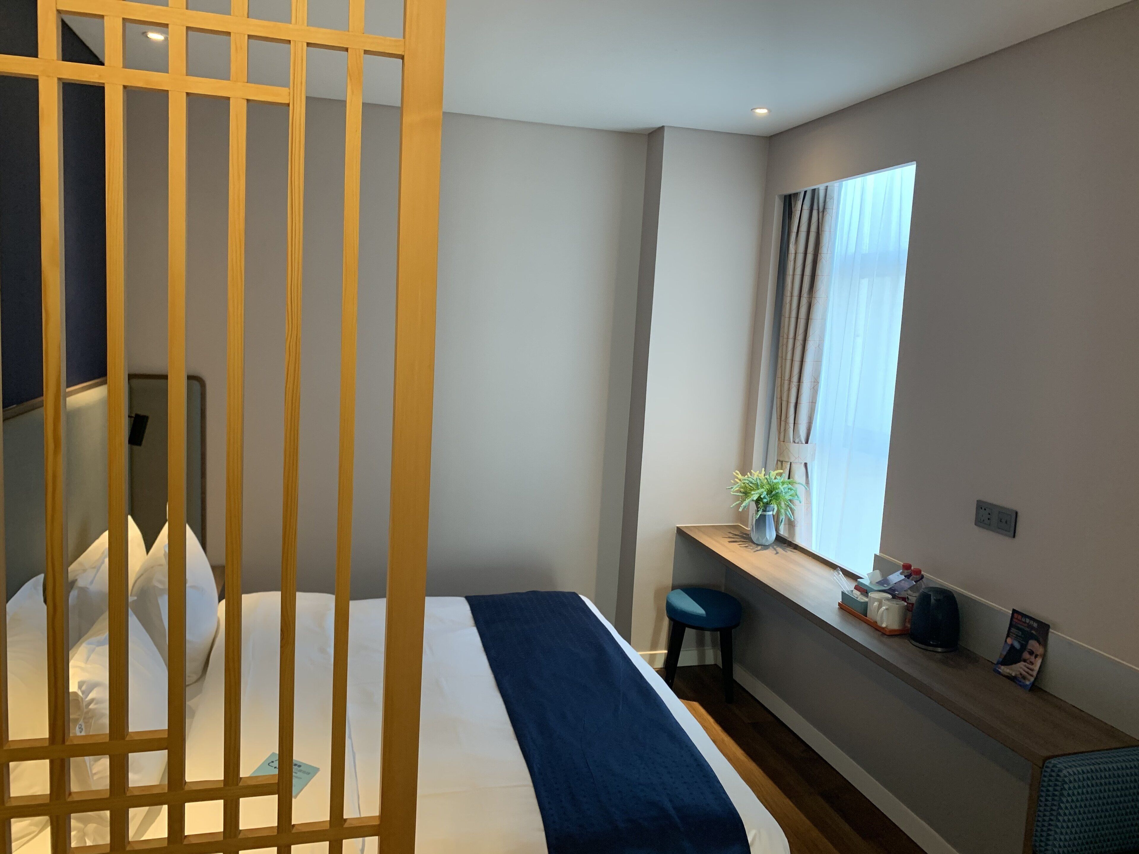 A hotel room with a bed, a desk, a window with curtains, and a small plant on the desk.