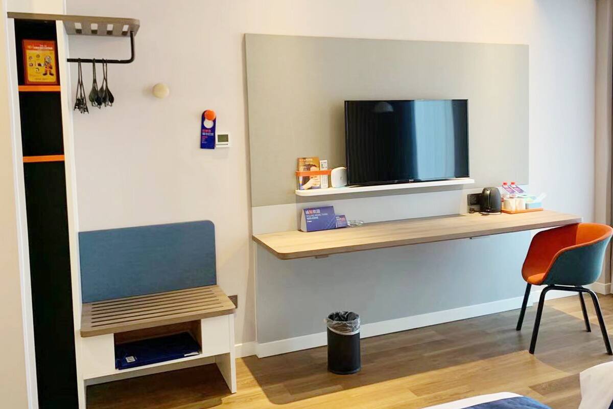 A hotel room with a wooden desk, a television, a blue chair, a small trash bin, and a bed.
