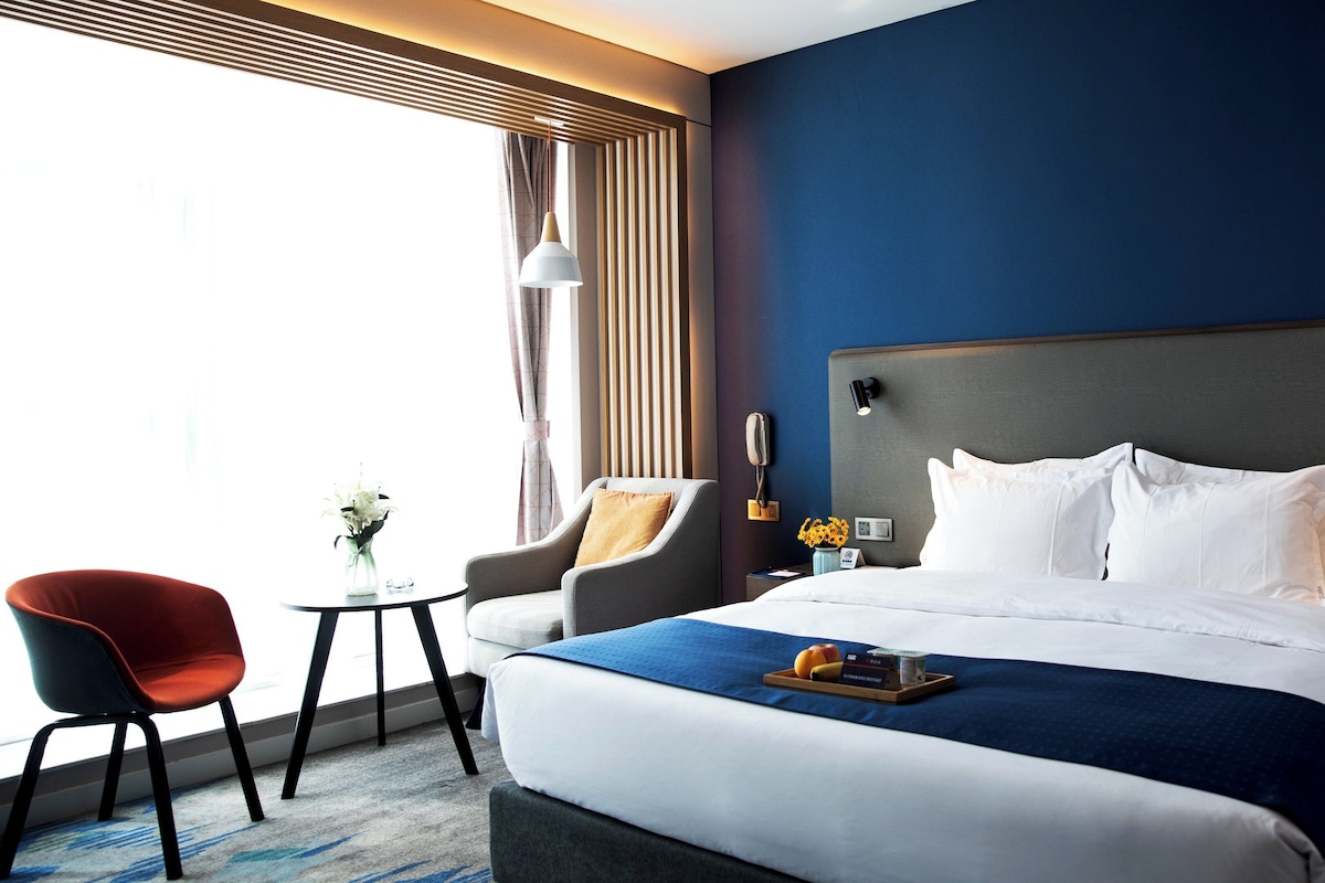 A hotel room with a large bed, a desk, a chair, and a window with curtains.