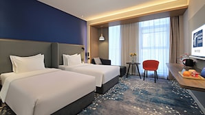 A hotel room with two beds, a desk, a chair, a small table with a vase, and a TV on the wall.